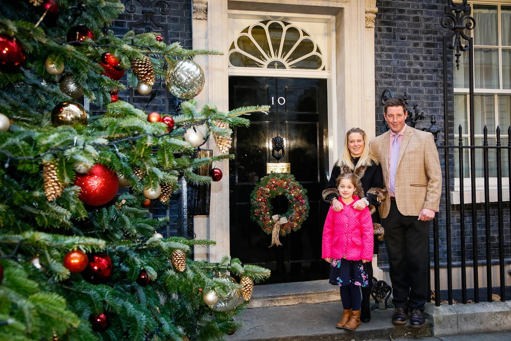 A family of three stands on the front steps of a 10 Downing Street, with a decorated Christmas tree on the left and a black door with a wreath behind them. The woman wears a black coat, the man wears a beige blazer, and a young girl in a bright pink jacket stands in the center.